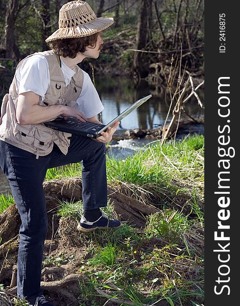 Man with portable computer standing and looking for something near small river at nature.