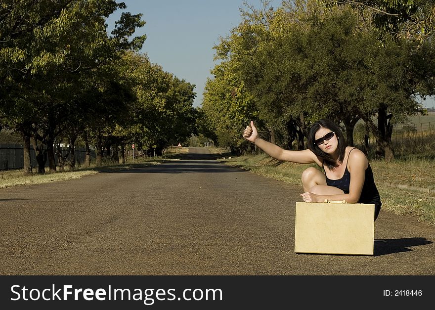 Brunette waiting roadside for a lift resting on suitcase with thumb in the air. Brunette waiting roadside for a lift resting on suitcase with thumb in the air