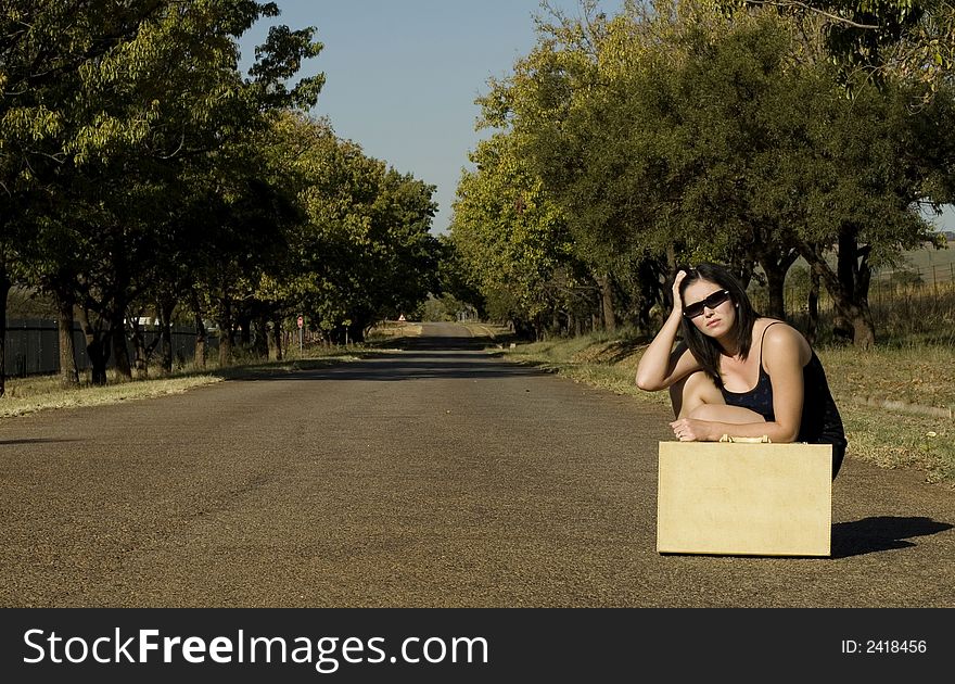 Brunette waiting roadside for a lift resting on suitcase with hands in hair. Brunette waiting roadside for a lift resting on suitcase with hands in hair