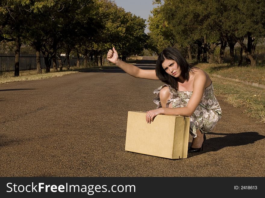 Brunette waiting roadside for a lift resting on suitcase with thumb in the air. Brunette waiting roadside for a lift resting on suitcase with thumb in the air