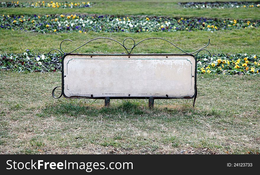 Empty information table in a park surrounded by grass and flowers behind it.