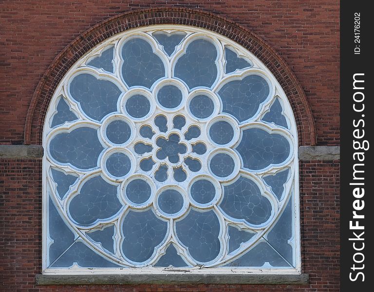 Old arched stained glass window with blue glass and white wood frame. Inside pattern is in petals and circles of tens. Exterior view against a brick wall. Old arched stained glass window with blue glass and white wood frame. Inside pattern is in petals and circles of tens. Exterior view against a brick wall.