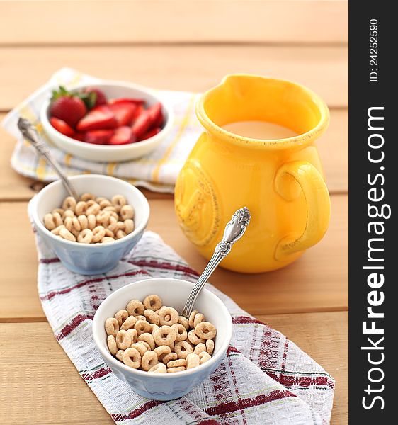 Close-up of a two bowls of cereal with a pitcher of fresh milk and strawberries.  Served on a wooden table, with a metal spoon on a napkin. Close-up of a two bowls of cereal with a pitcher of fresh milk and strawberries.  Served on a wooden table, with a metal spoon on a napkin.