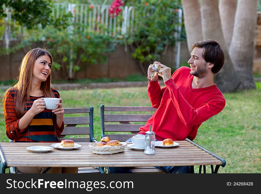 Smiling men photographing his girlfriend or wife while enjoying coffee and cakes at an outdoor cafe. Smiling men photographing his girlfriend or wife while enjoying coffee and cakes at an outdoor cafe