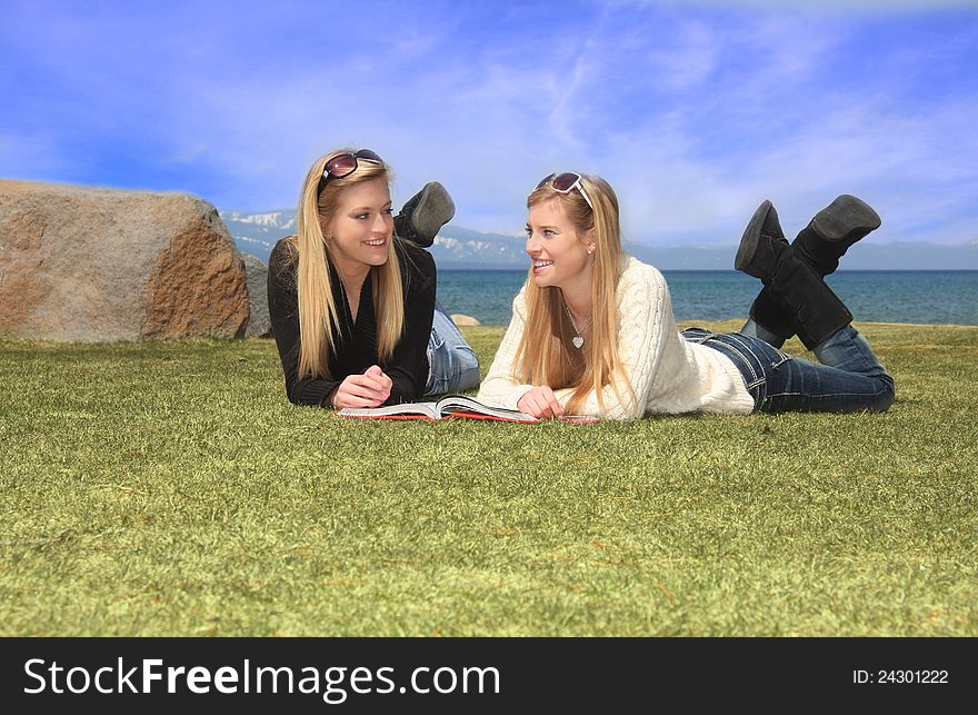 Two pretty teen aged girls laying on grass in front of a lake, studing from a book. Two pretty teen aged girls laying on grass in front of a lake, studing from a book.