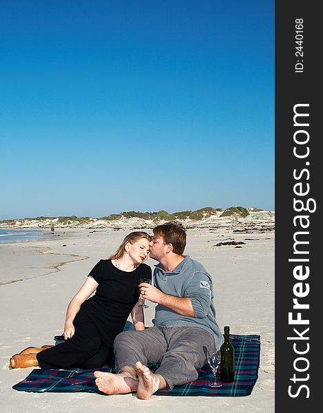 Young married couple sitting on the white sandy beach on their winter vacation in South Africa. They are enjoying the wine under the blue sky.