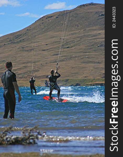 A kite surfer watching 3 other kitesurfers out in the water kitesurfing. All in wetsuits with scenic mountain behind. A kite surfer watching 3 other kitesurfers out in the water kitesurfing. All in wetsuits with scenic mountain behind
