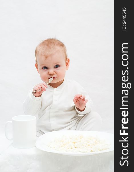 Cute little girl sitting on white blanket with spoon in her hands. There are white plate with curd and white cup in front of her. Cute little girl sitting on white blanket with spoon in her hands. There are white plate with curd and white cup in front of her.