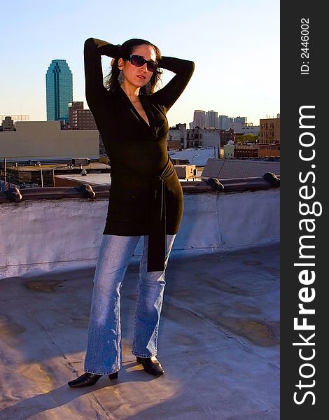 A woman posing as a fashion model in a black long shirt and jeans. She's wearing sunglasses and is holding up two arms. There's a skyscraper in the background.