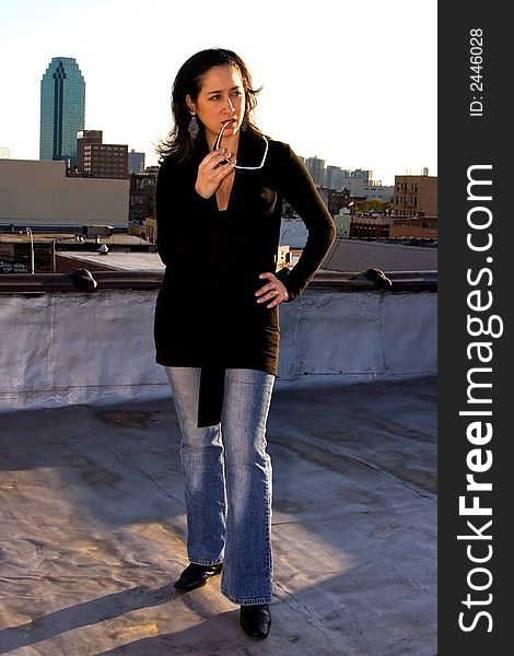 A woman posing as a fashion model in a black long shirt and jeans. She's holding sunglasses and chewing on them, with one end in her mouth. There's a skyscraper in the background. A woman posing as a fashion model in a black long shirt and jeans. She's holding sunglasses and chewing on them, with one end in her mouth. There's a skyscraper in the background.