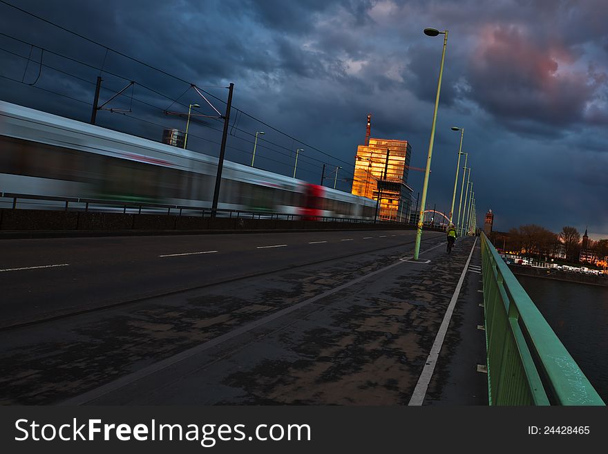 Tram crossing a bridge in Cologne, Germany. A tower building on the other side of the river rhine is illuminated by the setting sun and there is a dramatic clouded sky