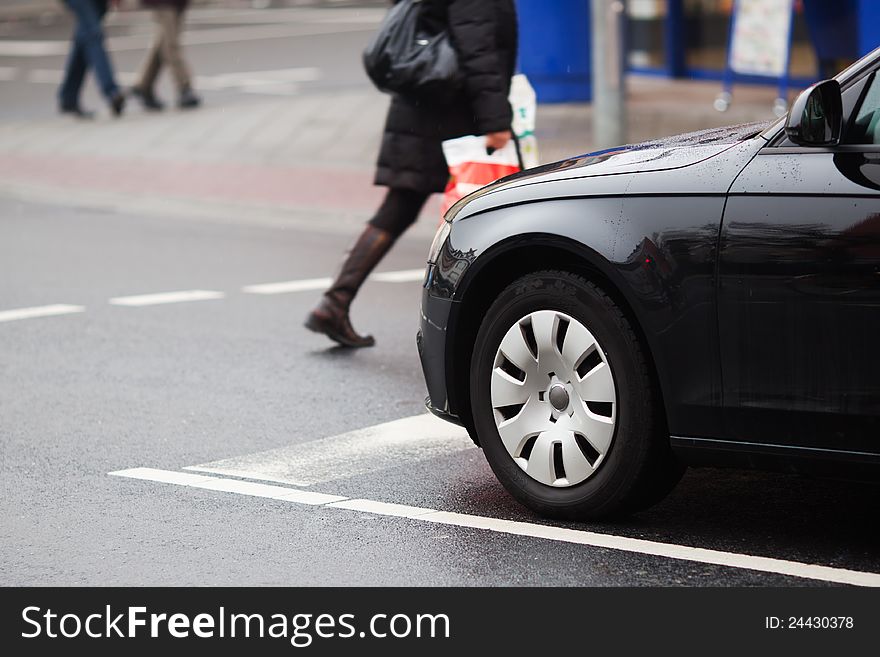 At a pedestrian crossing a woman crosses the street while a car waits at the stop line. At a pedestrian crossing a woman crosses the street while a car waits at the stop line