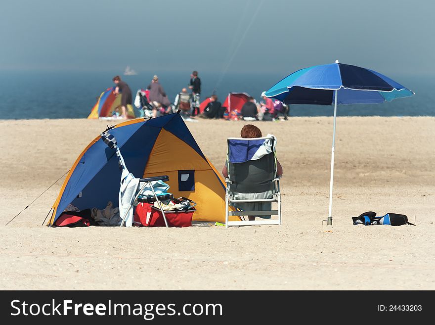 Scene at the beach with a unrecognizable person sitting on a deckchair, with a tent and a sunshade and other people in the farer background