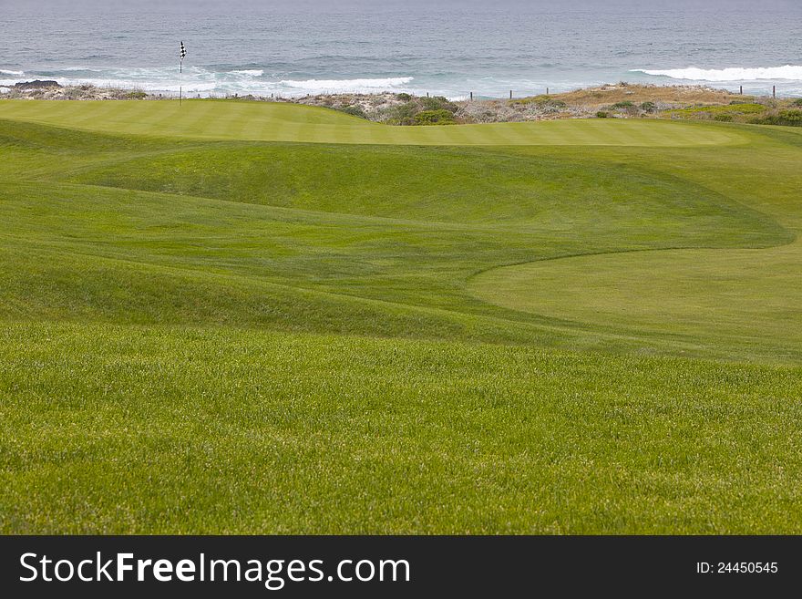 Beautiful, manicured golf course greens on a softly rolling hilly topography leading to the hole. The putting surface is located right by the ocean, with small waves washing into the shore. Beautiful, manicured golf course greens on a softly rolling hilly topography leading to the hole. The putting surface is located right by the ocean, with small waves washing into the shore.