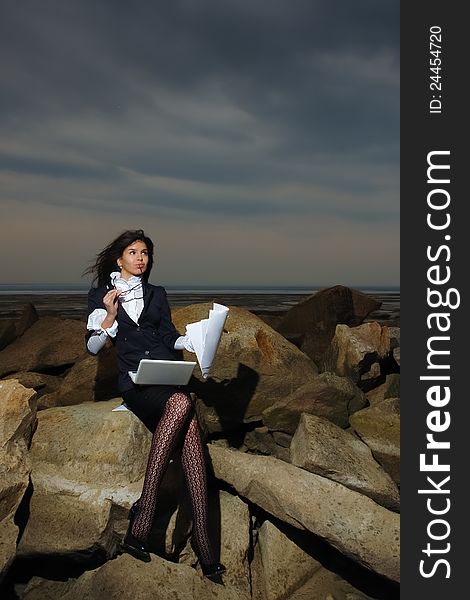 Business lady sitting on the rocks by the sea, against the backdrop of a cloudy sky. In the hands of her notebook and paper. Wind waving her hair.