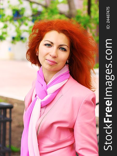 Outdoor portrait of a young smiling pretty red haired woman in pink jacket with a scarf on. Outdoor portrait of a young smiling pretty red haired woman in pink jacket with a scarf on