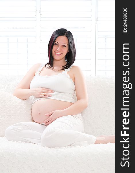 Portrait of a pretty young pregnant woman holding her belly. She is sitting on sofa and looking at camera. Portrait of a pretty young pregnant woman holding her belly. She is sitting on sofa and looking at camera.