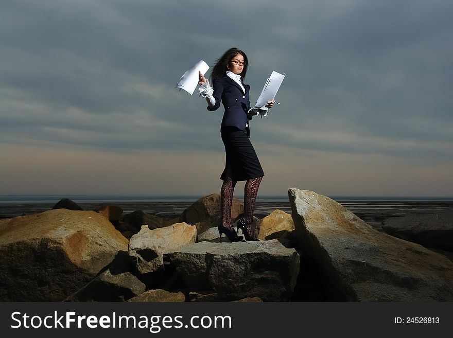 Business ladies standing on rocks by the sea, against the backdrop of a cloudy sky. In the hands of her paper. Wind waving her hair.
