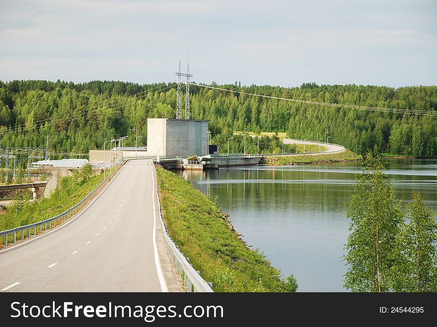 A hydroelectric power station is situated on the wide swift river. An asphalt road  passes by it and recedes into the taiga forest. A hydroelectric power station is situated on the wide swift river. An asphalt road  passes by it and recedes into the taiga forest.