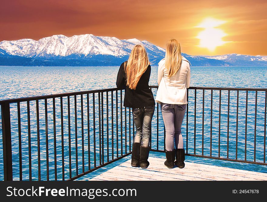 Two young women standing at the edge of a pier, over a lake, observing a beautiful sunset over snowcapped mountains. Two young women standing at the edge of a pier, over a lake, observing a beautiful sunset over snowcapped mountains.