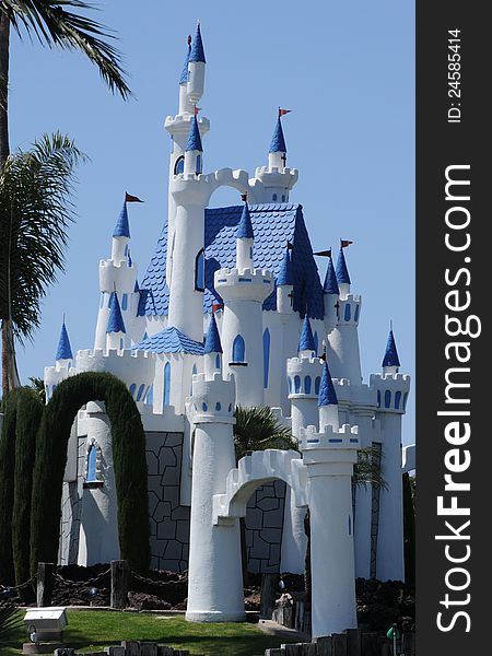 A fantasy castle in bright white wth blue trim, a brillant blue sky, green palm tree and some hedges border it