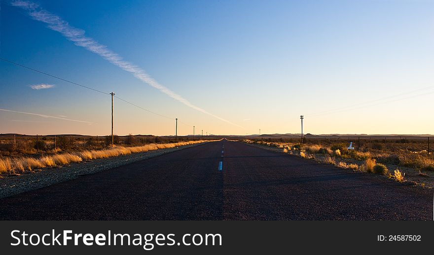 Empty highway in the arid Karoo region of South Africa at sunset bordered by telephone lines with vapour trails in the big sky. Empty highway in the arid Karoo region of South Africa at sunset bordered by telephone lines with vapour trails in the big sky.