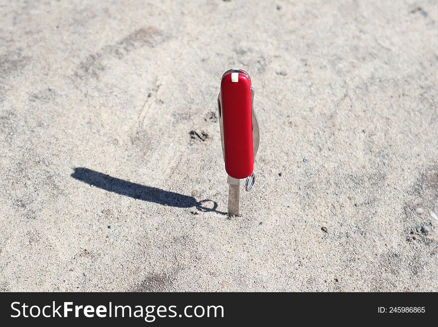 a red penknife stuck in the ground, sand