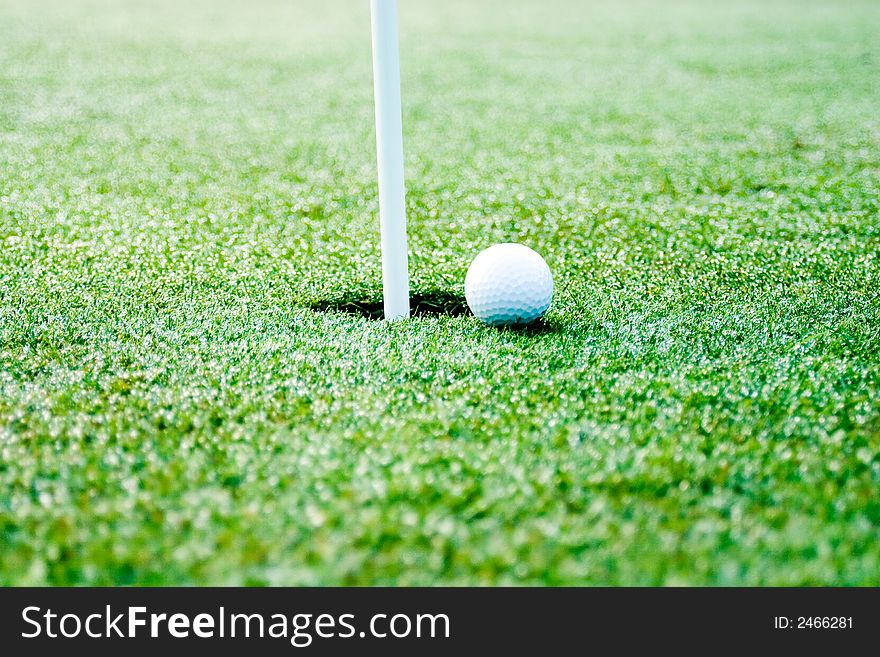 This is a picture of a golf ball next to the hole with flag pole showing. slective focus is on the ball and hole. This is a picture of a golf ball next to the hole with flag pole showing. slective focus is on the ball and hole.