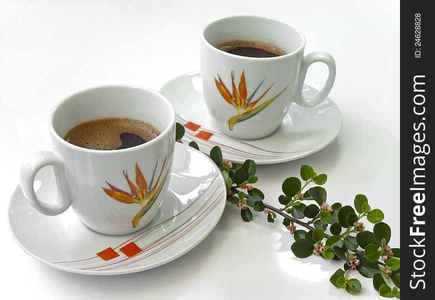 Two cups of coffee on a white table, with a floral sprig. Two cups of coffee on a white table, with a floral sprig