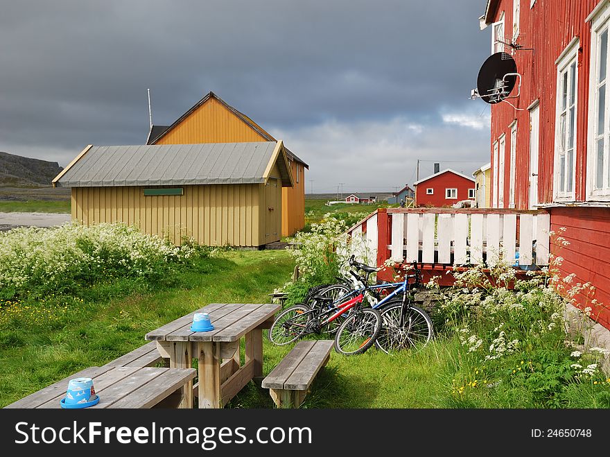 Norwegian fishing village is photographed in summer. Hamningberg is the easternmost point of Europe. There are several wood houses in the middle of the green grass under the overcast sky. Two bicycles are leaned against a wooden porch of the local cafe. Norwegian fishing village is photographed in summer. Hamningberg is the easternmost point of Europe. There are several wood houses in the middle of the green grass under the overcast sky. Two bicycles are leaned against a wooden porch of the local cafe.