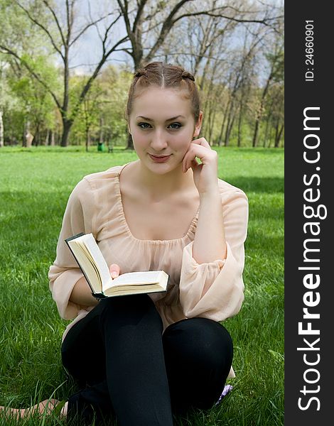 Young nice attentive woman lies on green grass and reads book against city park.