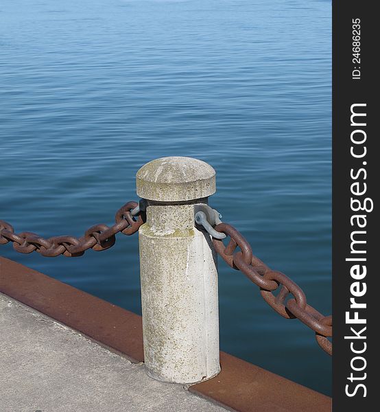 Concrete post and heavy chain along the edge of a pier, with blue water in the background. Concrete post and heavy chain along the edge of a pier, with blue water in the background.