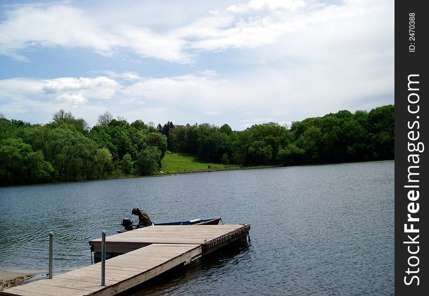 A man in a boat who can be seen from the dock, on a beautiful lake with a distant park.