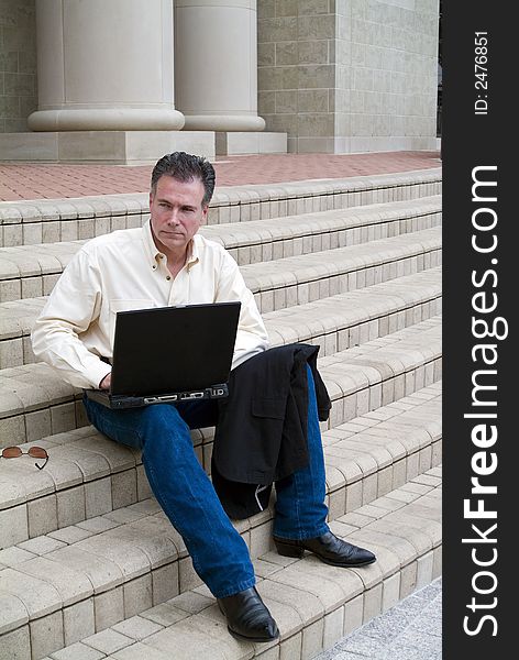 Man sitting on the steps of a large building with a laptop with an expression of contemplation. Man sitting on the steps of a large building with a laptop with an expression of contemplation.