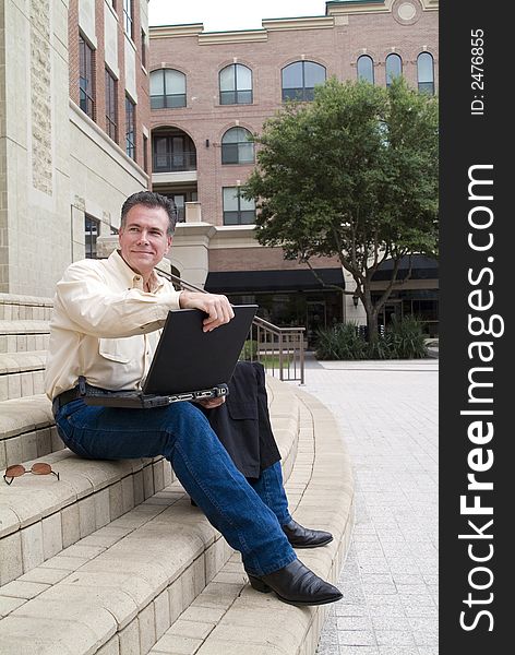 Man sitting on the steps of a large building with a laptop and a friendly little smile on his face. Man sitting on the steps of a large building with a laptop and a friendly little smile on his face.