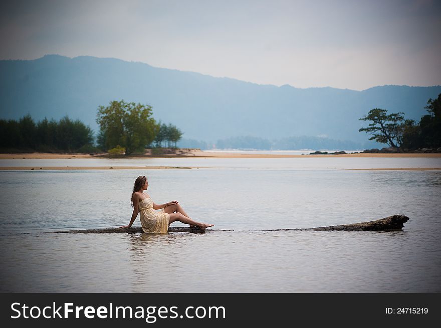 A beautiful young girl in white dress sitting on a log in the water. A beautiful young girl in white dress sitting on a log in the water