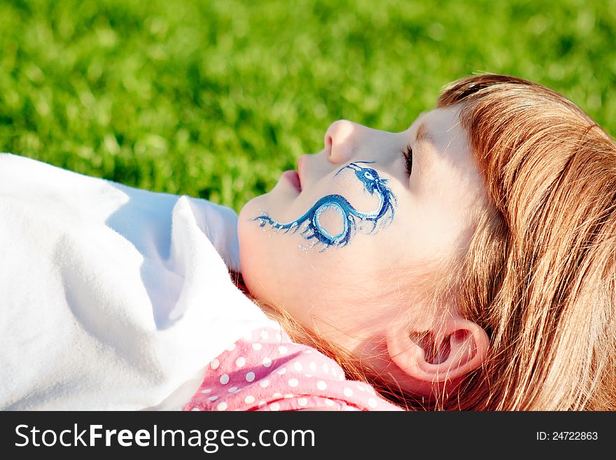 Portrait of a young girl with blue dragon on her face lying on a lawn