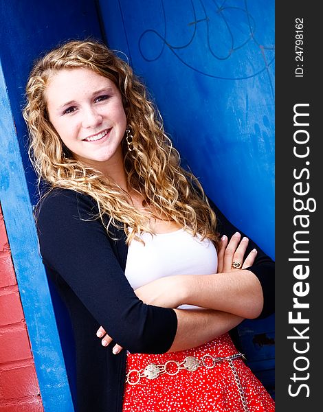 A beautiful blond teenage girl next door with curly hair and a pretty smile in a red skirt, white top, black sweater leaning against a blue door with her arms crossed in an urban setting. A beautiful blond teenage girl next door with curly hair and a pretty smile in a red skirt, white top, black sweater leaning against a blue door with her arms crossed in an urban setting.