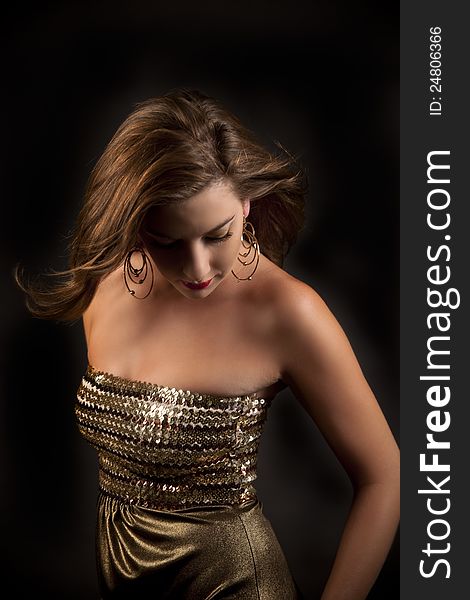 Beautiful caucasian woman in gold dress, gold earrings and dramatic makeup looking down with her hair blowing in the breeze against black background. Beautiful caucasian woman in gold dress, gold earrings and dramatic makeup looking down with her hair blowing in the breeze against black background.