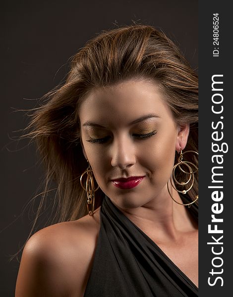 Beautiful caucasian woman in dramatic golden makeup with dark lashes and large golden drop earrings looking down while her long brown hair blows around her face against a dark background. Beautiful caucasian woman in dramatic golden makeup with dark lashes and large golden drop earrings looking down while her long brown hair blows around her face against a dark background