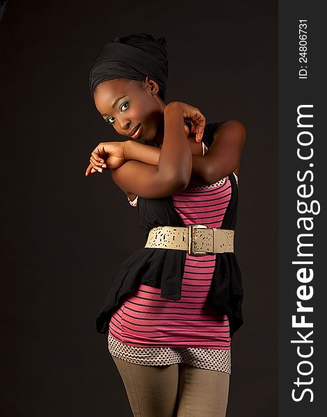 Beautiful african woman posing in fashionable casual street wear consisting of beige belt, pink shirt, pants, black jacket and headband, glancing at viewer against black background. Beautiful african woman posing in fashionable casual street wear consisting of beige belt, pink shirt, pants, black jacket and headband, glancing at viewer against black background