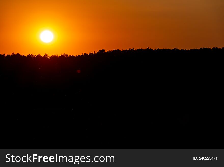 Beautiful sunset over the forest on a summer day. The dark silhouette of the forest and the bright blinding orange sun lightly touches the treetops on the horizon