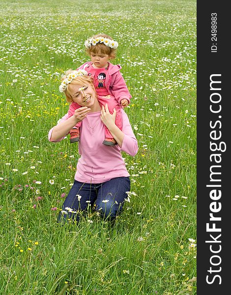 Baby girl sitting on her mothers neck wearing daisy wreath and picking more daisies on a meadow full of flowers. Baby girl sitting on her mothers neck wearing daisy wreath and picking more daisies on a meadow full of flowers