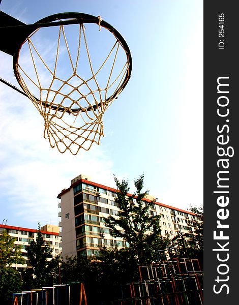 Basketball hoop in school grounds with apartment buildings, trees and playground equipment in the background