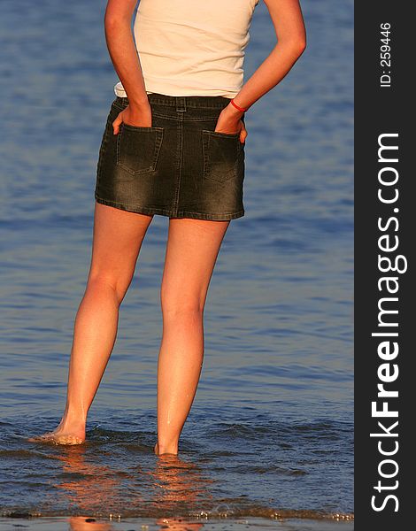 A woman in a short denim skirt paddling in the sea, with her hands in her pockets..