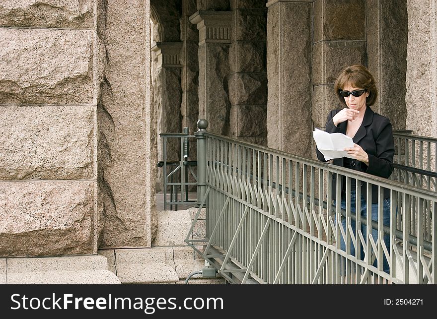 A woman standing on a ramp leading to a large building reading something on a few sheet of paper she is holding in her hand. A woman standing on a ramp leading to a large building reading something on a few sheet of paper she is holding in her hand.