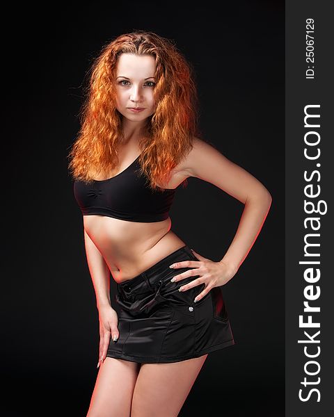 Portrait of a sexual redhead girl in a black bra and black leather skirt on a dark background