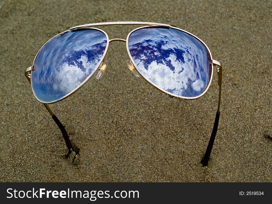 Close up of sunglasses on the sand gggreflecting a cloudy sky. Close up of sunglasses on the sand gggreflecting a cloudy sky