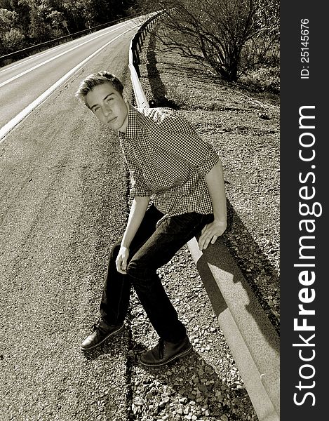 A nostalgic looking photo of a young man along the side of the road sitting on the gaurd rail. A nostalgic looking photo of a young man along the side of the road sitting on the gaurd rail