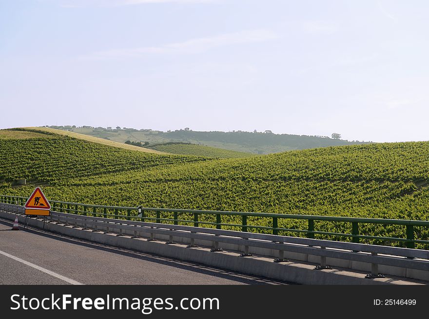 Warning sign of works on a highway, passing throw a small mountain planted with vines.